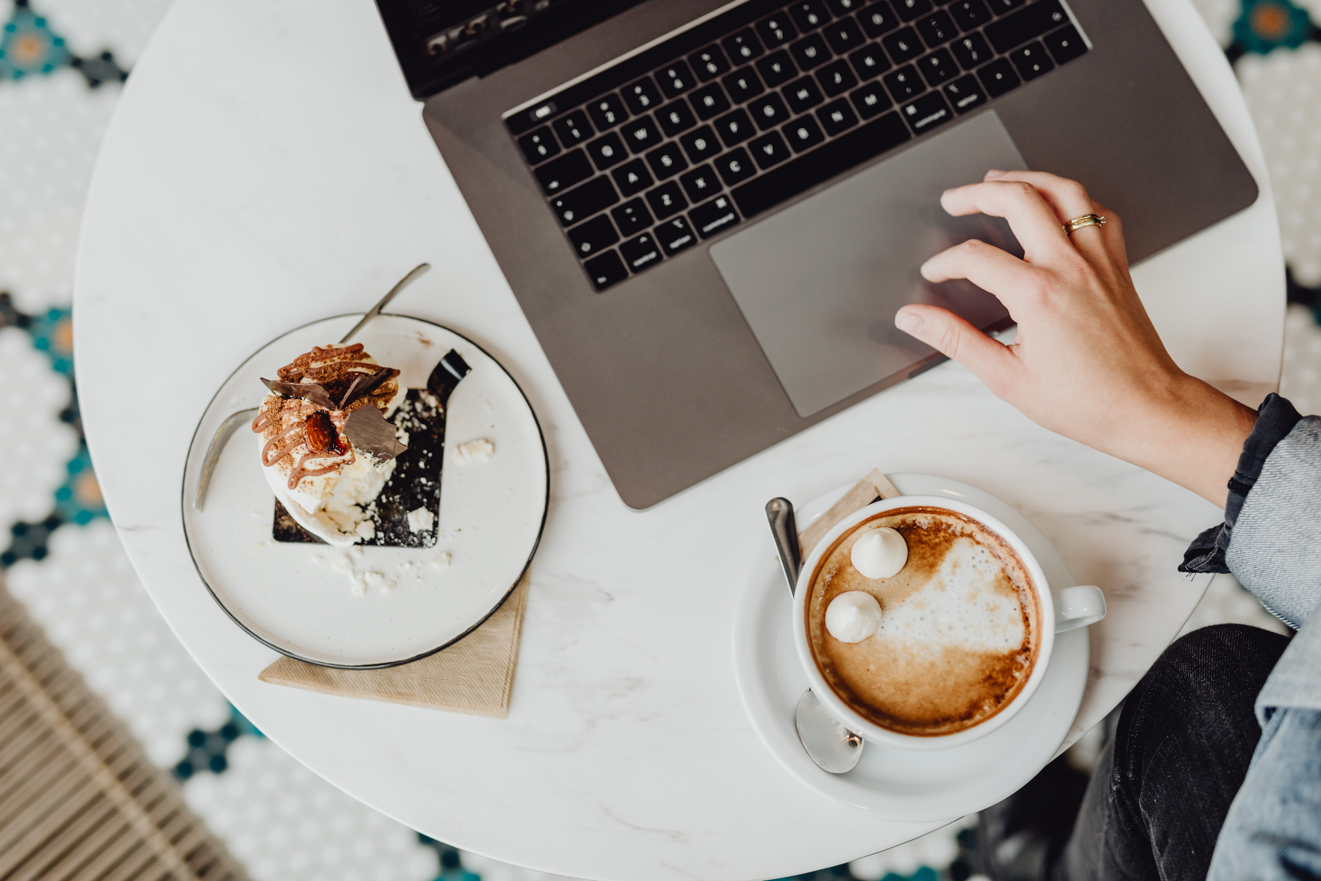 Overhead view of a bright white marble cafe table with an open laptop, a cappuccino with latte art, and a plated dessert
