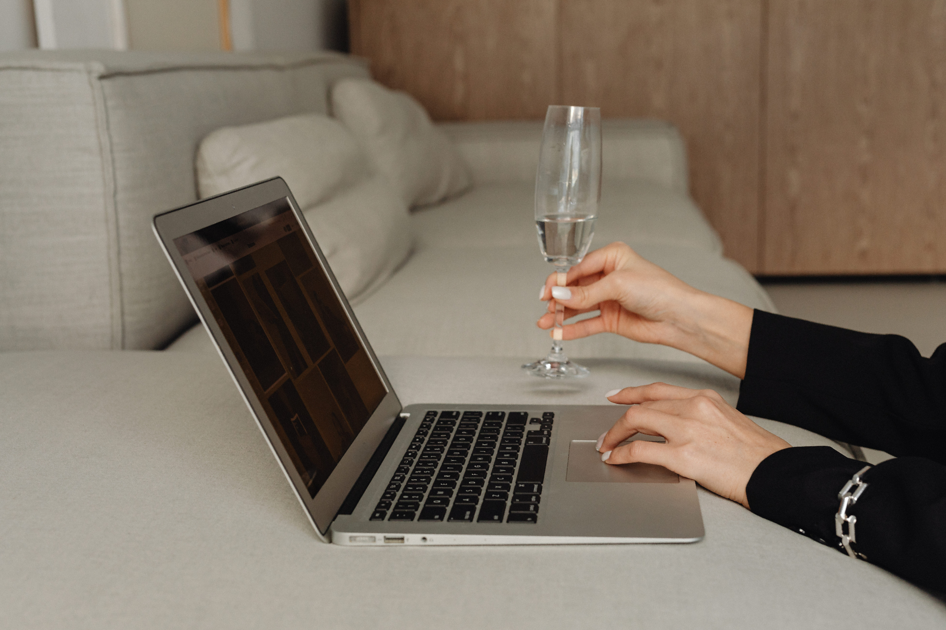 Woman in a black blazer working on a laptop on a light couch while holding a champagne flute