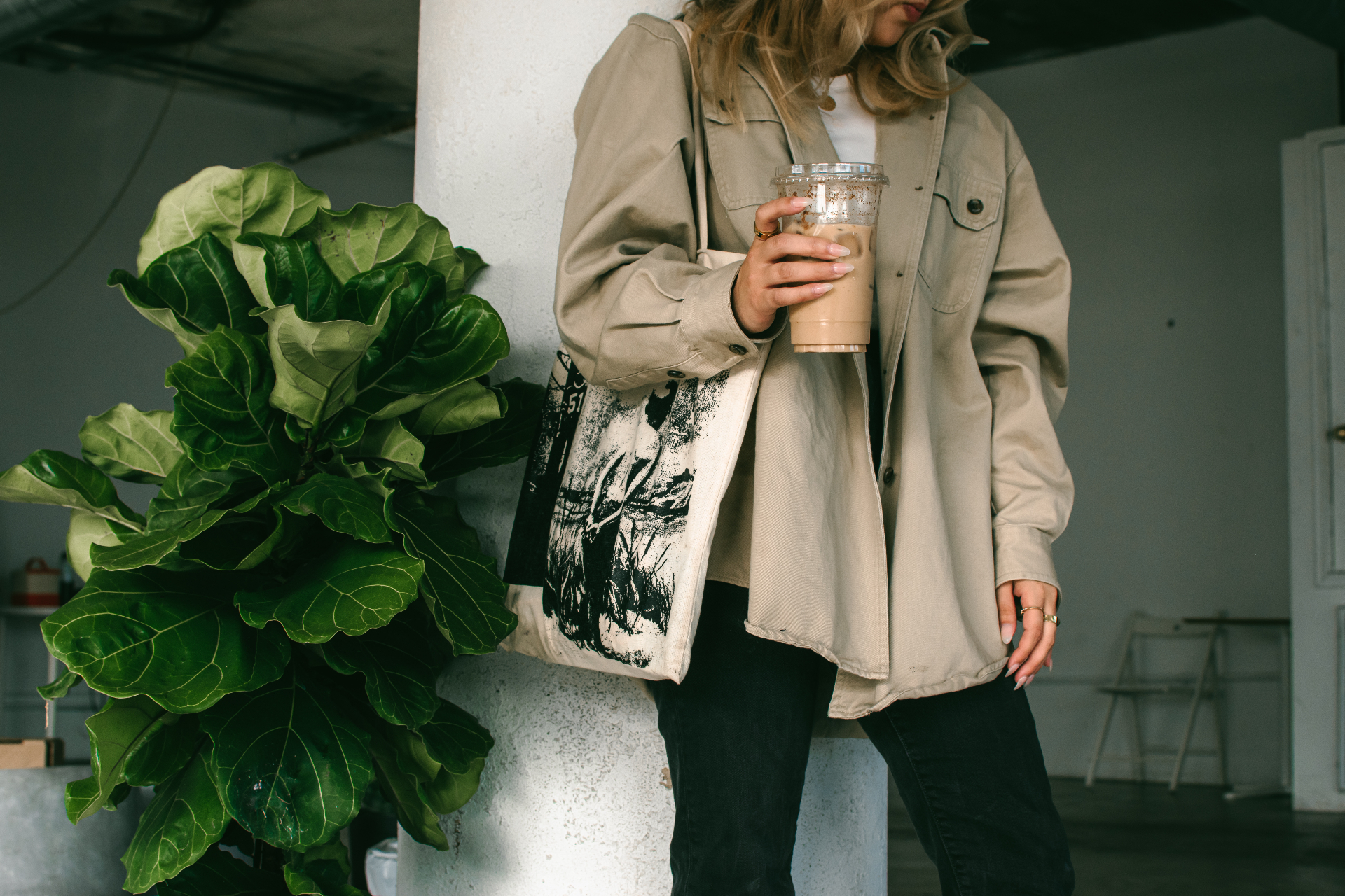 Woman in a beige overshirt holding an iced coffee beside a large fiddle-leaf fig plant in a softly lit studio space