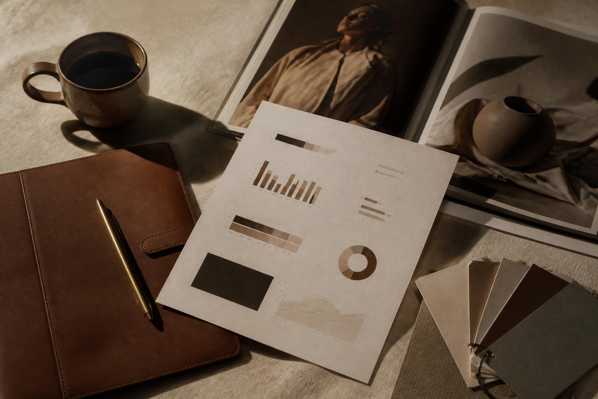 Overhead flatlay of a coffee mug, reading glasses, a green spiral notebook, a phone showing a calendar app, and a grey textured pouch on a wood desk