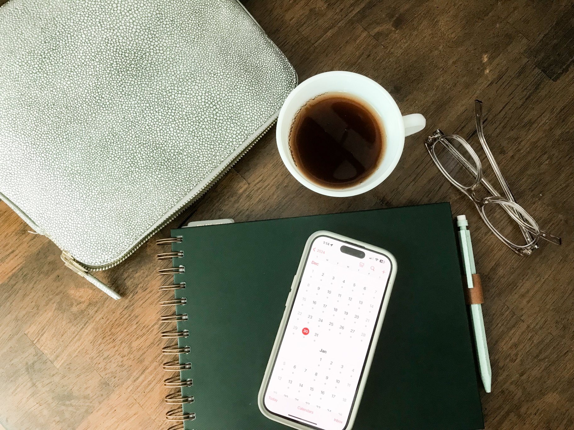 Overhead flatlay of a coffee mug, reading glasses, a green spiral notebook, a phone showing a calendar app, and a grey textured pouch on a wood desk